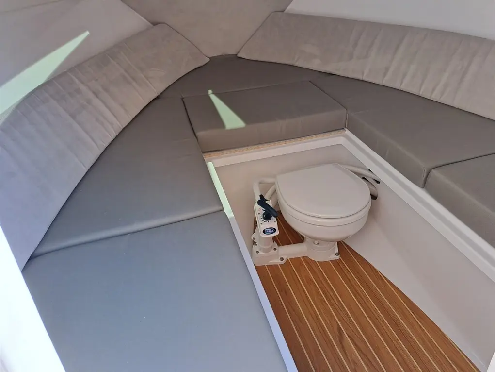 Interior of a boat featuring gray cushioned seating, a wooden floor, and a compact marine toilet in a well-lit space.